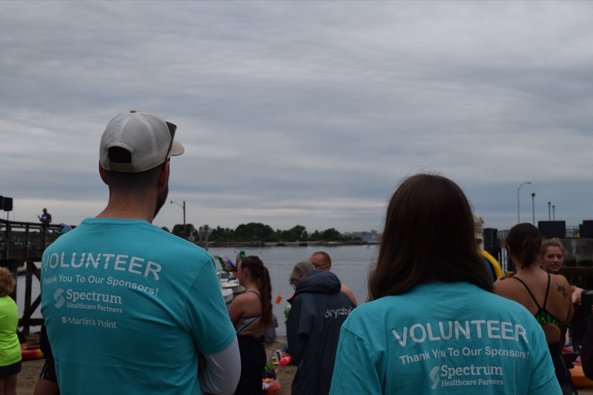 Two people in volunteer shirts, seen from behind, watching swimmers on the beach